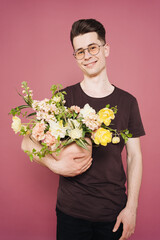 A young man is holding a beautiful bouquet of flowers in his hands on a pink background. The guy decided to make a surprise to his girlfriend. Mother's Day. Women's day. Flowers delivery.