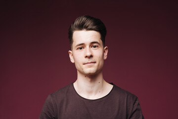 Fototapeta premium Isolated shot of young serious handsome male with trendy hairdo, wears casual brown t shirt, has serious expression as listens to interlocutor, poses in studio against crimson background