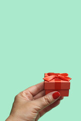 Mão de uma pessoa segurando um presente pequeno, embrulhado com papel e laço vermelhos com fundo verde claro.