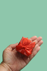 Mão de uma pessoa segurando um presente pequeno, embrulhado com papel e laço vermelhos com fundo verde claro.