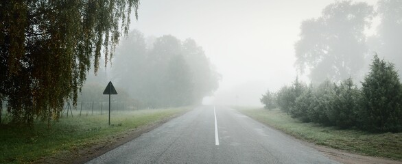 An empty highway (asphalt road) through the fields and forest in a thick fog at sunrise....