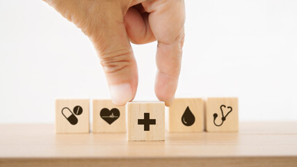 senior's hand holding hospital or clinic symbol on wood cube with other blurred health and medical icon for health insurance, wellness, wellbeing concept