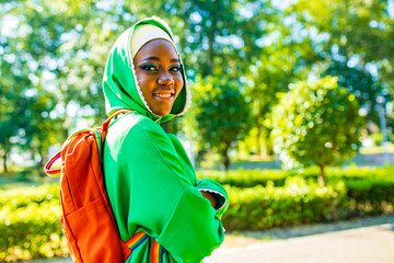 african woman in green hijab with bright makeup with rainbow backpack in summer park