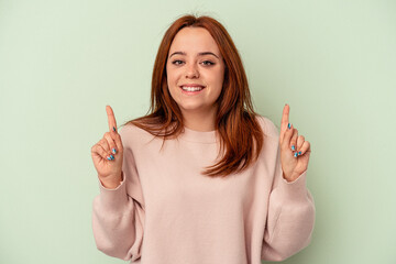 Young caucasian woman isolated on green background indicates with both fore fingers up showing a...