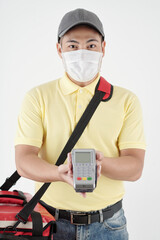 Fototapeta na wymiar Positive young delivery man in medical mask ready to receive payment giving payment terminal to customer
