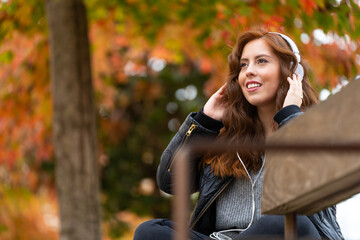 Fototapeta na wymiar Girl listening to music with white headphones on a park bench in autumn