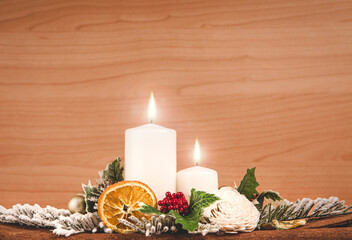 Christmas decorative composition with lighted white candles, oranges, berries, white flowers, pine branches, leaves on cinnamon sheet and wooden background