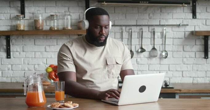 Bearded african american man freelancer in headphones eats cookies and works at the computer while sitting at the kitchen table at home.