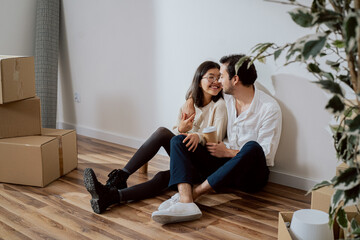 A couple in love are sitting by a wall in a new apartment, a gallant man dressed in a white shirt embraces a smiling pretty woman, they are drinking coffee, flirting, around unpacked boxes of things