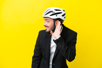 Business caucasian man with a bike helmet isolated on yellow background listening to something by putting hand on the ear