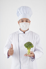 Portrait of positive chef in protective mask holding fresh broccoli and showing thumbs-up