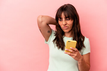 Young Argentinian woman holding a mobile phone isolated on pink background touching back of head, thinking and making a choice.