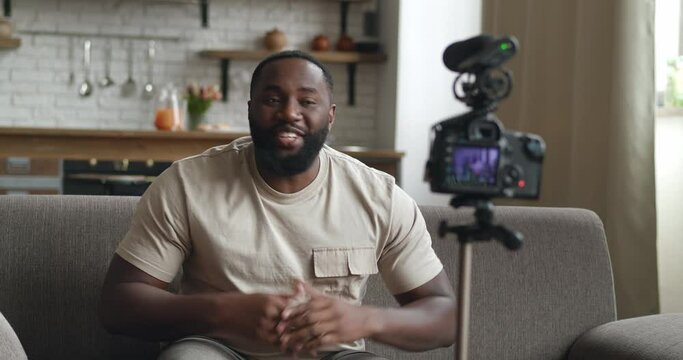 Excited african man blogger filming vlog video for internet with professional camera at home. Male content creator sitting on sofa and filming online lessons. Vlogger recording vlog with videocamera