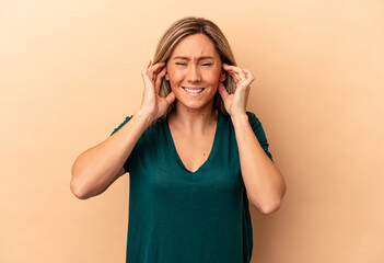 Young caucasian woman isolated on beige background covering ears with hands.