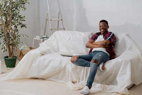 A Happy, Dark-skinned Man Sits On A Couch Covered With Plastic Sheeting In New Apartment During Renovations, The Boy Is Proud Of The Move, Smiling With Arms Crossed Over His Chest