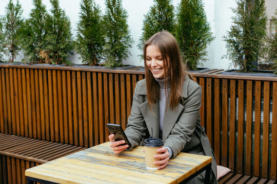 Beautiful smiling girl with glasses sitting in cafe, holding smartphone in hands and chating