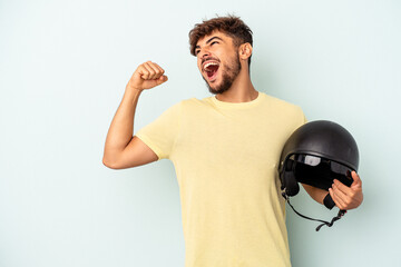 Young mixed race man holding motorcycle helmet isolated on blue background raising fist after a victory, winner concept.