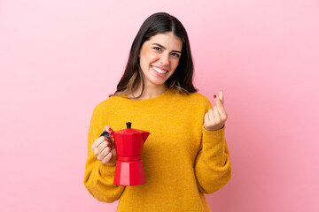 Young Italian woman holding a coffee maker isolated on pink background making money gesture