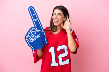 Young Italian fan woman with foam hand isolated on pink background listening to something by putting hand on the ear