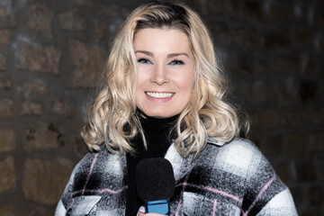 blonde reporter in coat and microphone smiling at the camera