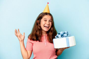 Little caucasian girl celebrating her birthday holding a cake isolated on blue background receiving a pleasant surprise, excited and raising hands.