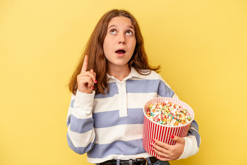 Little caucasian girl holding popcorns isolated on yellow background pointing upside with opened mouth.