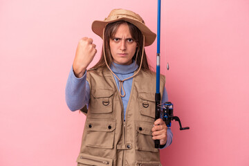 Young caucasian fisherwoman holding a rod isolated on pink background showing fist to camera, aggressive facial expression.