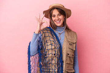 Young caucasian fisherwoman holding a net isolated on pink background cheerful and confident showing ok gesture.