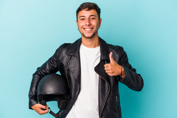 Young biker man holding helmet isolated on blue background  smiling and raising thumb up