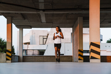 Black sportswoman listening music while running on parking