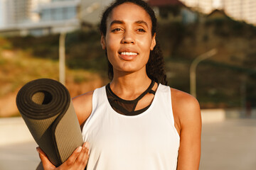 Black sportswoman smiling while standing with fitness mat on parking