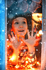 smiling sweet child girl standing by window at Christmas time. With colorful lights from Christmas...