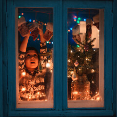 smiling sweet child girl standing by window at Christmas time. With colorful lights from Christmas...
