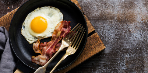 Traditional English breakfast with fried eggs and bacon in cast iron pan on dark concrete background. Top view.