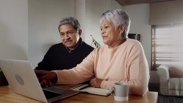 Elderly couple discussing a budget on laptop, sitting at kitchen counter. Pen and notebook in hand