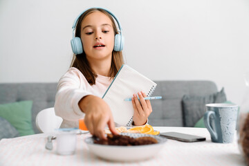 Naklejka premium White girl in headphones writing down notes while having breakfast