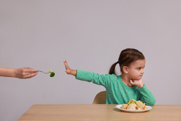 Cute little girl refusing to eat vegetables at table on grey background