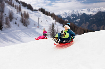 childhood, sledging and season concept - happy little kids sliding on sleds down snow hill in...