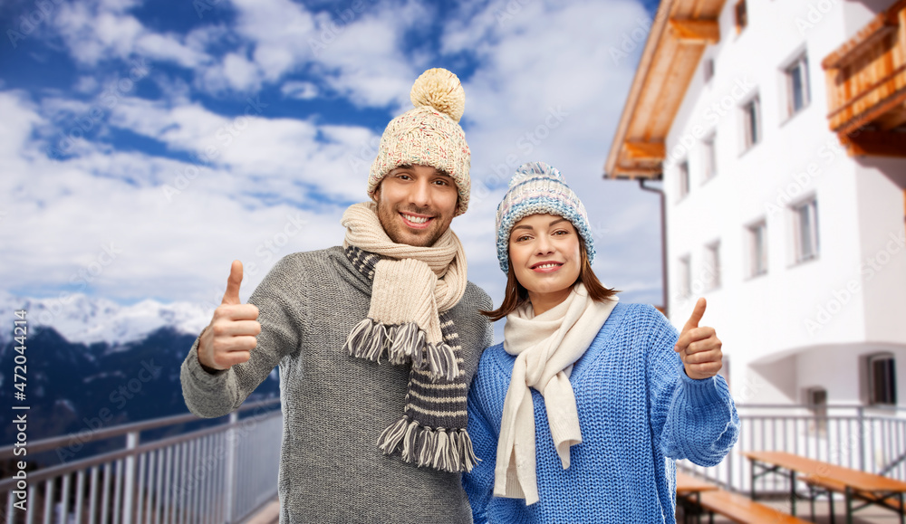 Poster travel, tourism and winter holidays concept - happy couple in knitted hats and scarves showing thumbs up over mountains and ski resort background