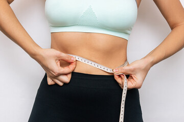 Cropped shot of a young slender woman in sports clothes measuring her waist with a centimeter isolated on a white background. Close-up. Diet, sport and wellness concept