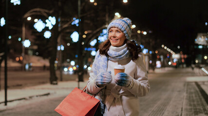 holidays, christmas and people concept - happy young woman with shopping bag drinking takeaway coffee and walking along city street in winter
