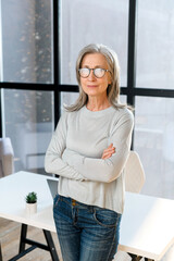 Confident and smiling old senior business woman wearing smart casual shirt and stylish eyeglasses stands with arms crossed in modern office space. A mature bossy lady, elderly purposeful leader