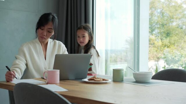 Pensive Asian mother working on laptop while her daughter asking for something in the morning