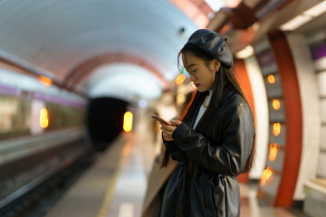 Korean girl with smartphone at empty metro platform. Stylish asian female browsing social media waiting for underground train. Young woman dependent from mobile phone and online communication concept