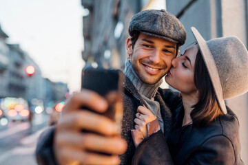 Young woman kissing happy boyfriend while taking selfie on smart phone