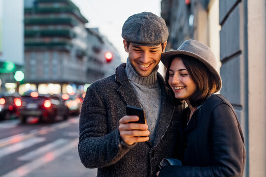 Happy couple in warm clothing sharing smart phone on street