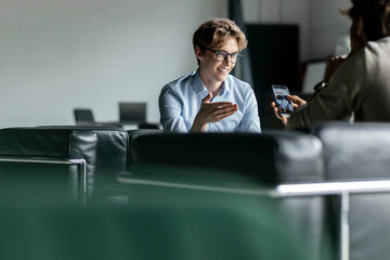 Smiling young businessman gesturing in front of colleague using mobile phone at lobby