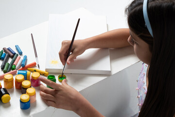 cute girl painting canvas, putting brush inside green paint, next to colored paints, photo seen from above.