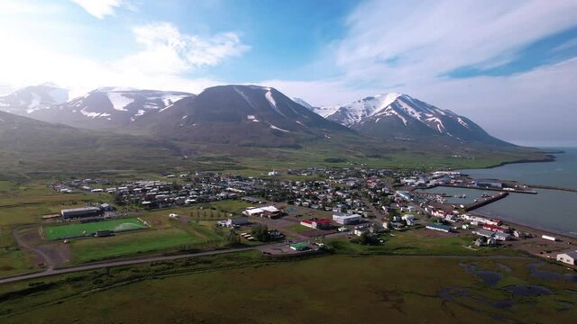 Beautiful turning aerial shot of coastal Icelandic town. Shot in summer with lots of green. Snowy mountains in distance. 