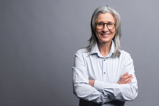 Successful senior mature gray-haired businesswoman with arms crossed isolated on gray, elder female entrepreneur ceo manager in glasses standing and confidently looking at the camera, hands folded
